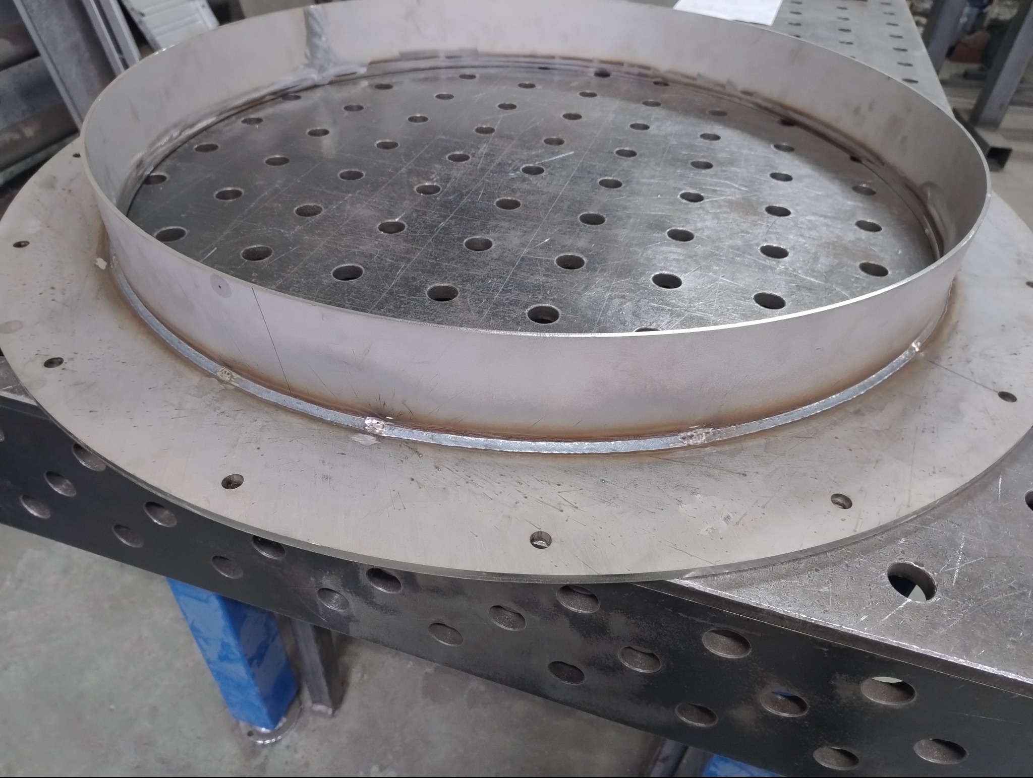 Szlifowany, okrągły element ze stali nierdzewnej po spawaniu, leżący na perforowanym stole spawalniczym. Widoczne ślady po spawaniu i obróbce.