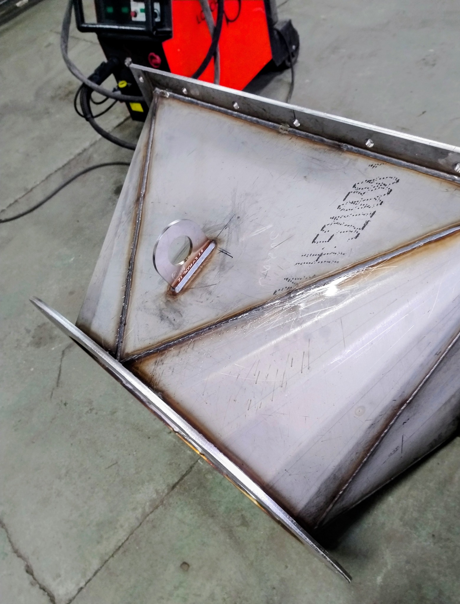 Spawana konstrukcja ze stali nierdzewnej z widocznymi śladami spawania i elementem mocującym. W tle spawarka.