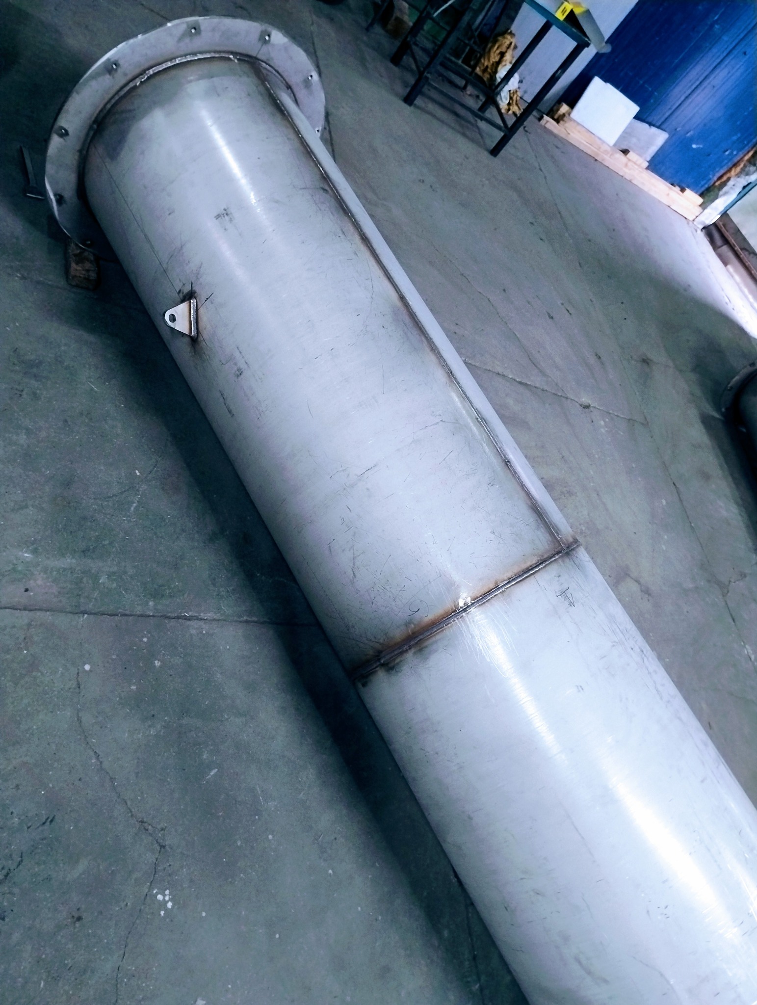 Spawana, długa rura ze stali nierdzewnej z kołnierzem, leżąca na szarym podłożu. Widoczne ślady spawania i mocowania. W tle fragment warsztatu.