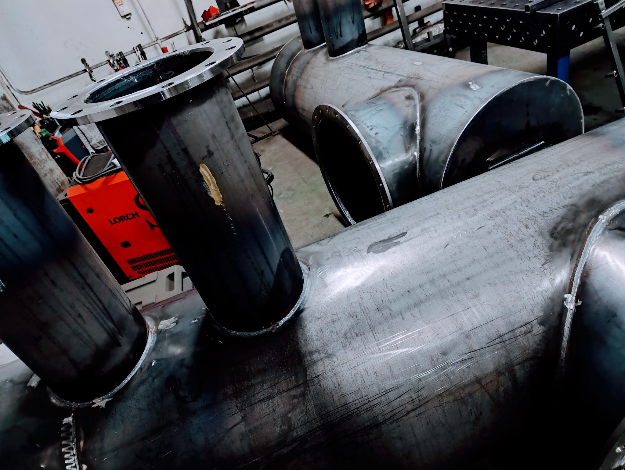 Konstrukcja ze spawanych rur stalowych z kołnierzami, widoczna spawarka Lorch w tle. Elementy połączone precyzyjnymi spawami, surowy metaliczny wygląd.