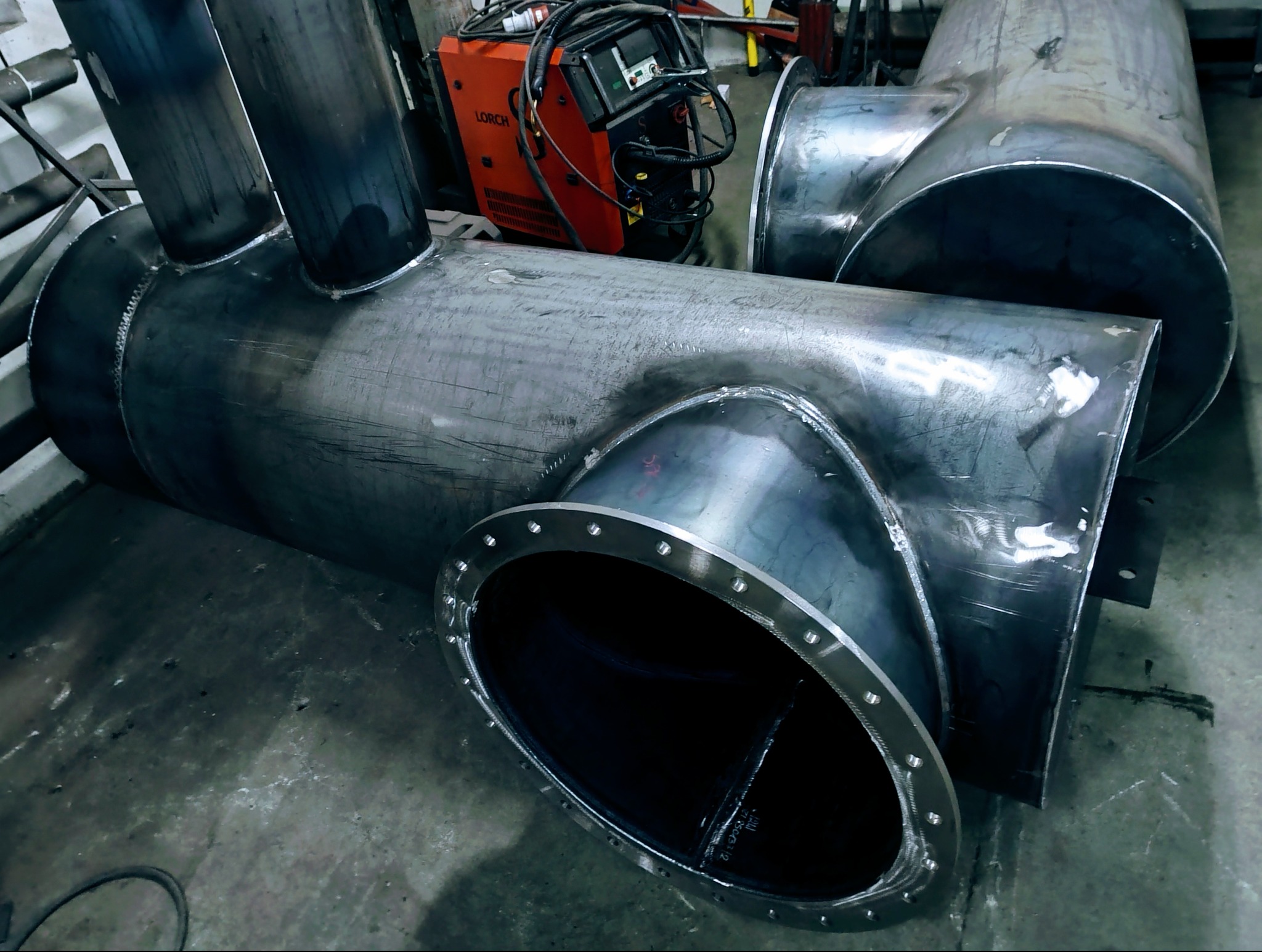 Spawane, metalowe rury z kołnierzem w Garbatce-Letnisko. Widoczny spawacz Lorch w tle. Elementy konstrukcyjne przed montażem, surowy metal z widocznymi śladami obróbki.