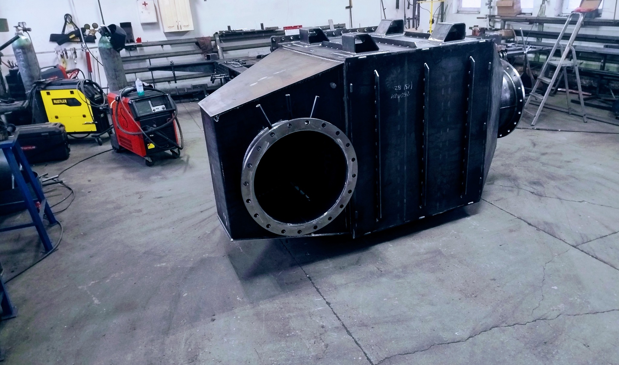 Duży, spawany, metalowy element konstrukcyjny z okrągłym otworem na flanszę, widoczny w warsztacie z narzędziami spawalniczymi w tle. Surowy, przemysłowy wygląd.