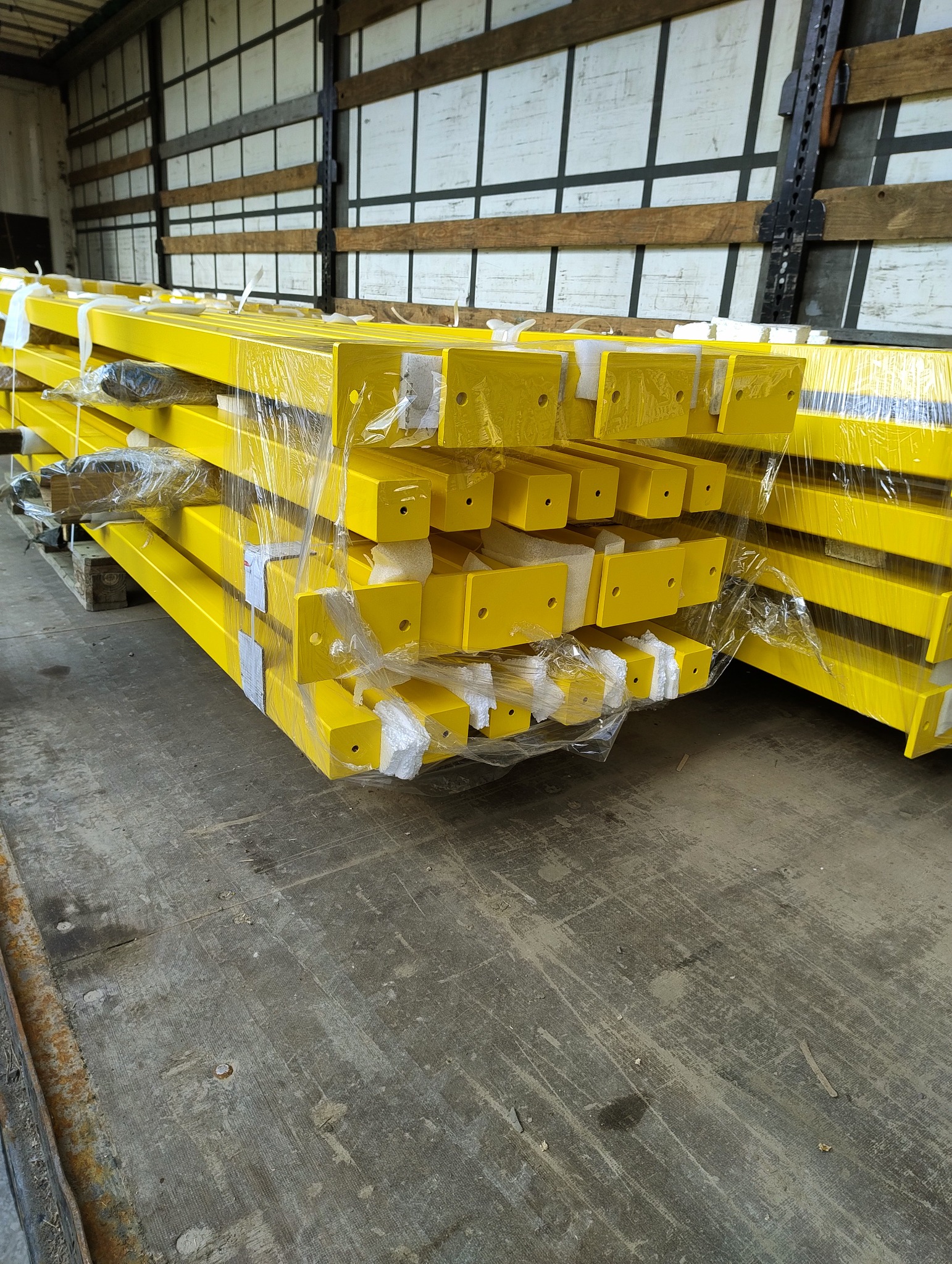 Żółte, metalowe profile, zabezpieczone folią, ułożone w stos na palecie wewnątrz pojazdu dostawczego. Widoczne otwory montażowe.