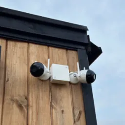 Dwie kamery monitoringu Dahua zamontowane na drewnianej ścianie budynku z czarnymi elementami wykończeniowymi, widok z dołu na tle pochmurnego nieba.