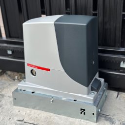 VGS Michał Kozłowski - Zamknięty automat do bram przesuwnych Nice Hi-Speed w kolorze szaro-czarnym, zamontowany na metalowej podstawie, widoczny na tle czarnego ogrodzenia.