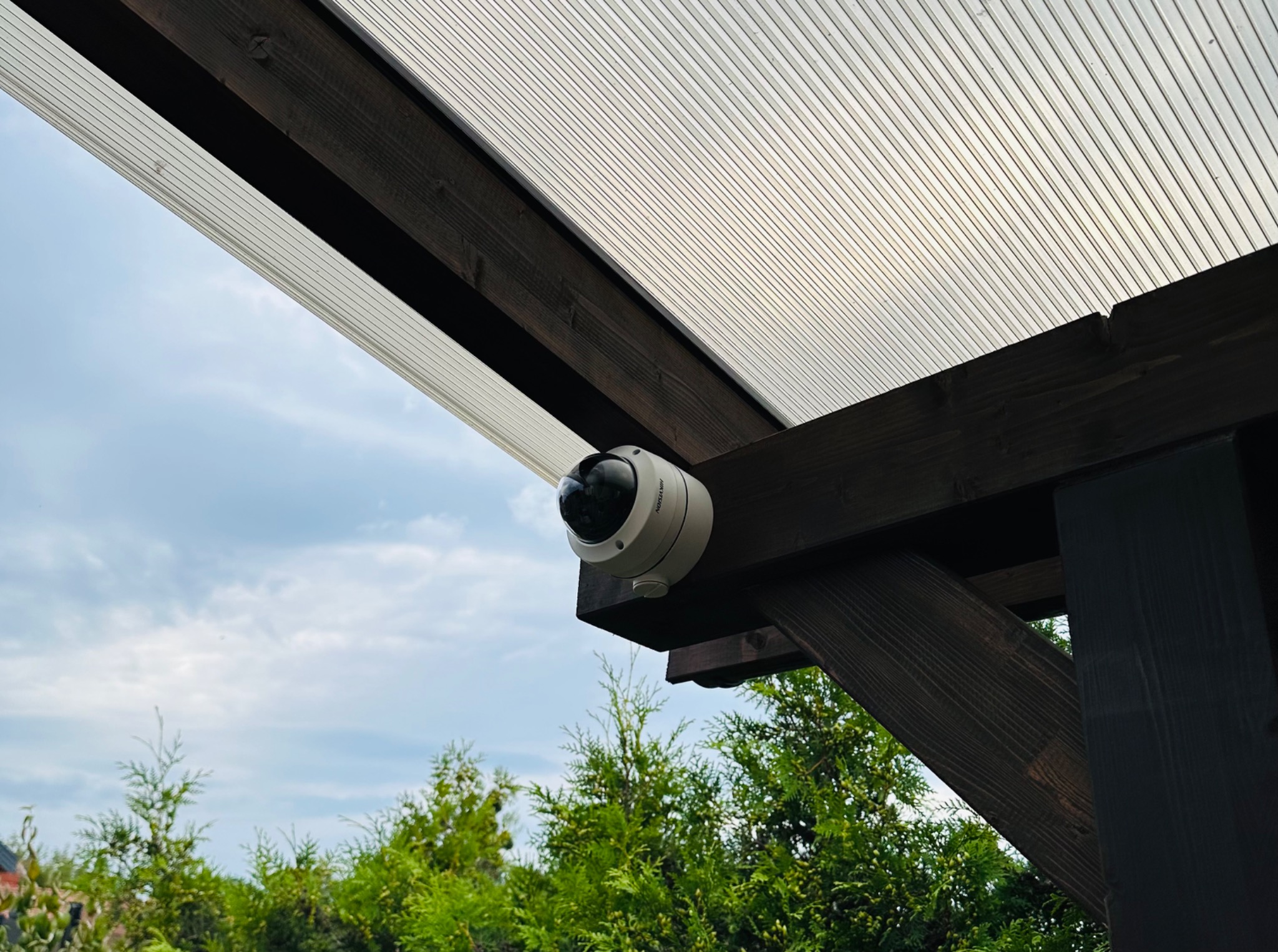Kamera monitoringu Hikvision zamontowana pod drewnianą konstrukcją z poliwęglanu w Olszewnicy Starej, widok z dołu na tle nieba i zieleni.