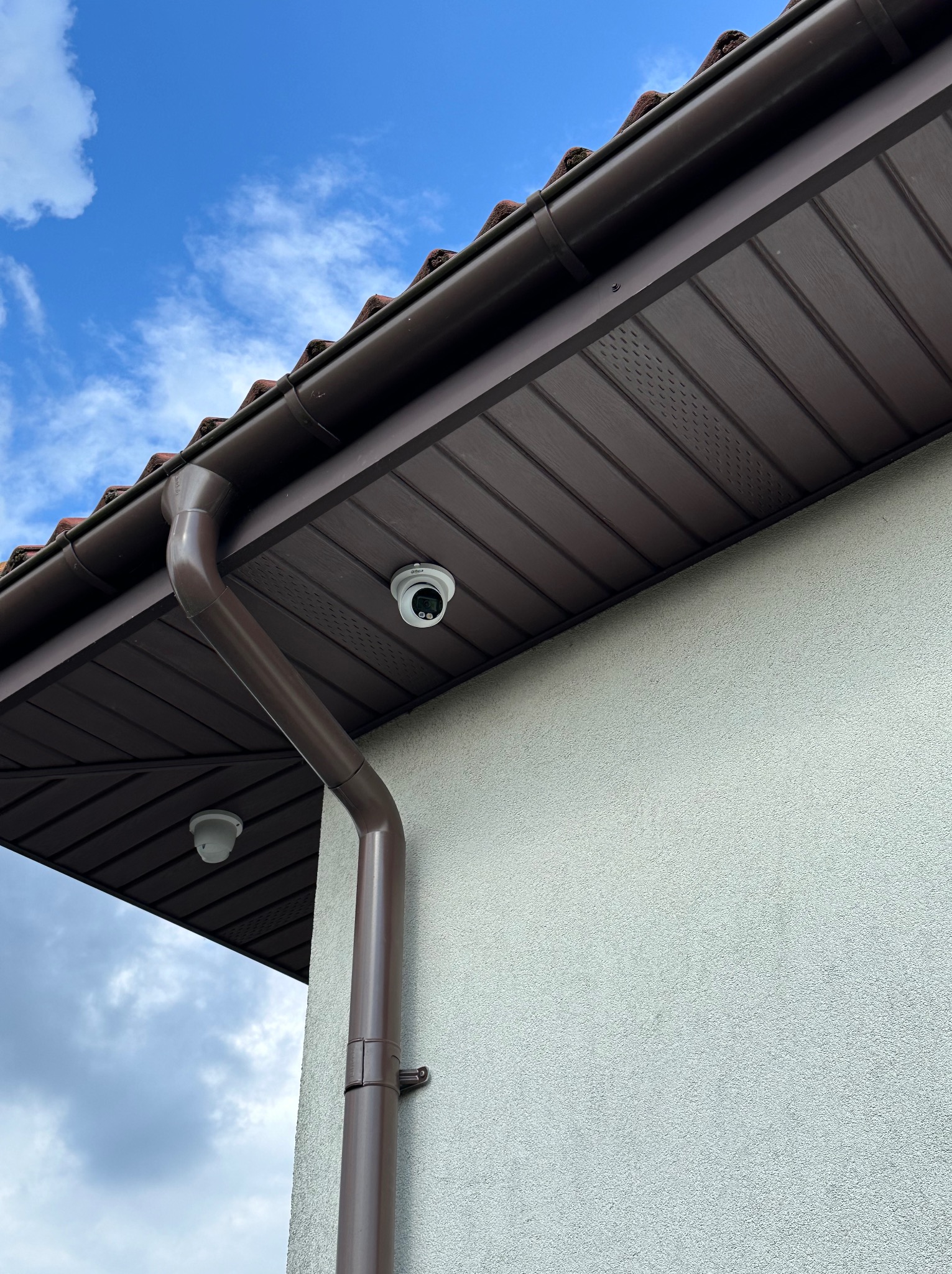 Dwie kamery monitoringu zamontowane pod okapem domu, widoczna brązowa rynna i fragment elewacji w jasnym kolorze.