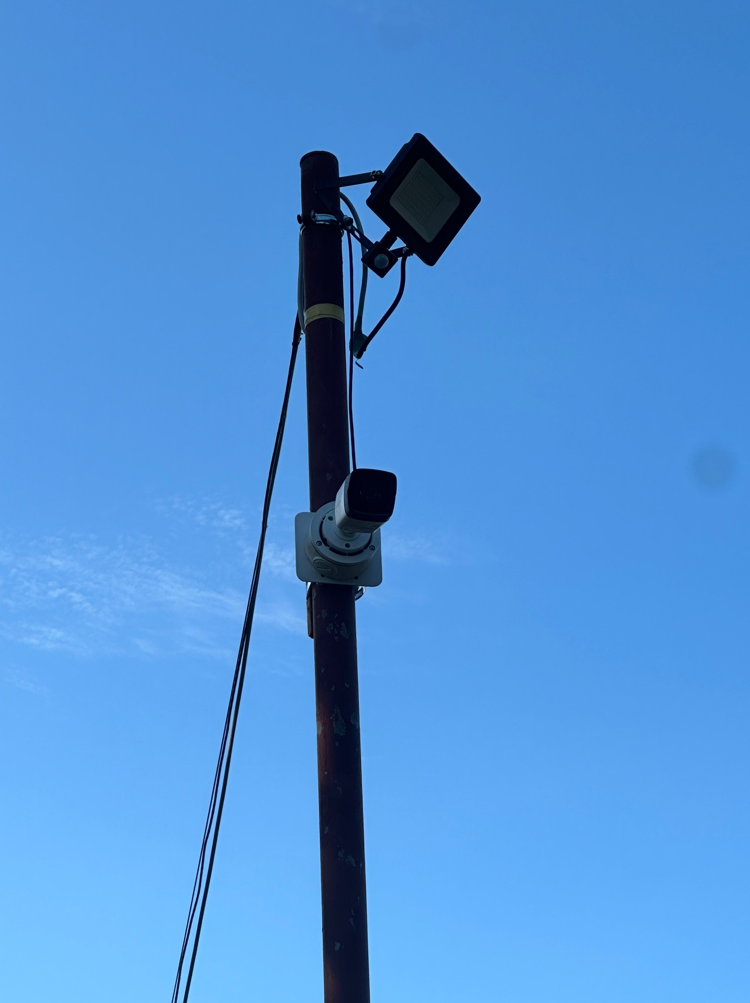 Słup z zamontowaną kamerą monitoringu i reflektorem skierowany w górę na tle błękitnego nieba.