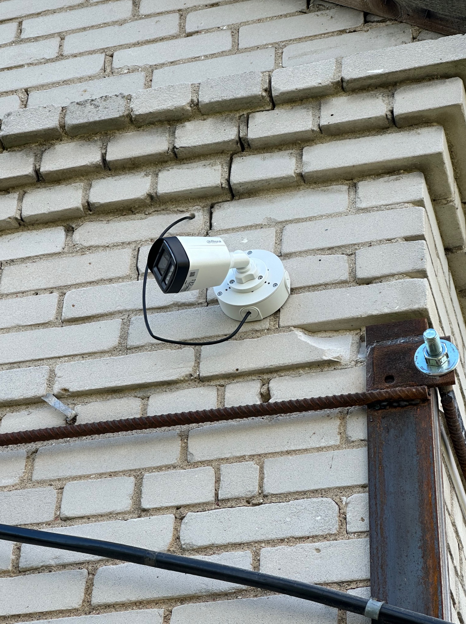 Kamera monitoringu Dahua zamontowana na białej ceglanej ścianie budynku, widoczne przewody i metalowa konstrukcja wsporcza.