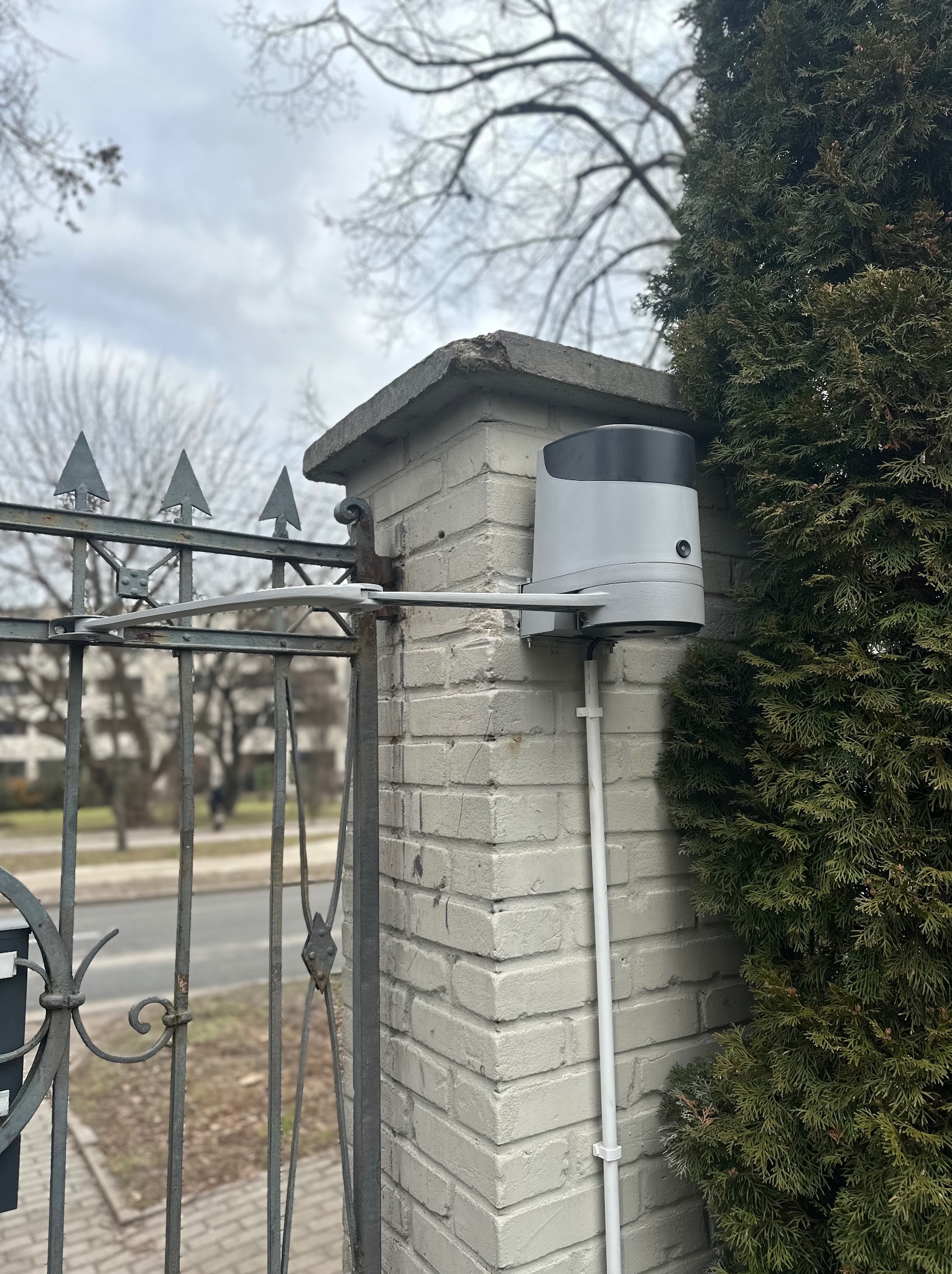 Szary automat do bramy skrzydłowej zamontowany na ceglanym słupku z widoczną metalową bramą i tujami w tle, uchwycony w pochmurny dzień.