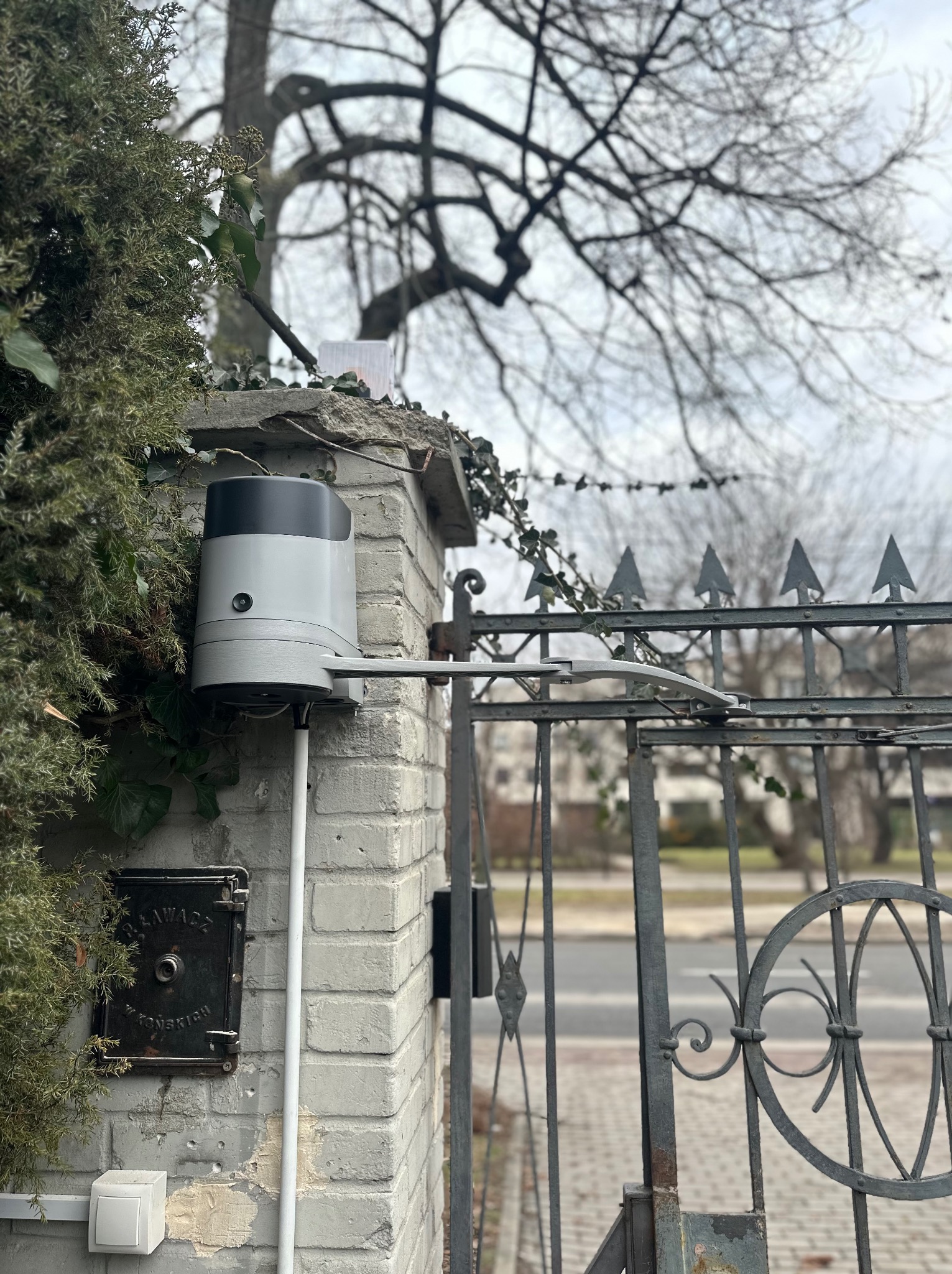 Szary napęd bramy zamontowany na ceglanym słupku z widocznym mechanizmem ramienia, brama metalowa z ozdobnymi elementami, zarośnięty murem bluszcz, pochmurne niebo w tle.