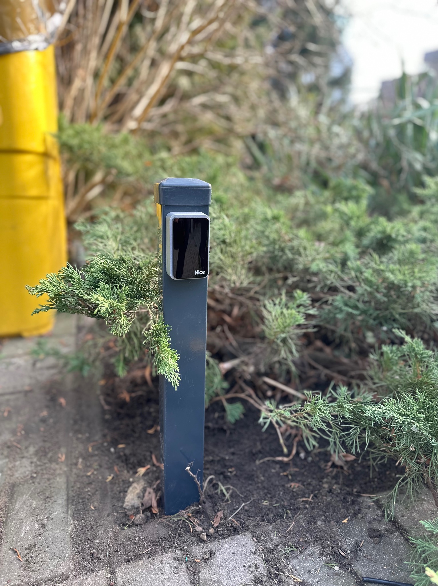Szary słupek z zamontowanym panelem sterującym bramą marki Nice, otoczony zielonymi krzewami, z żółtym pojemnikiem w tle.