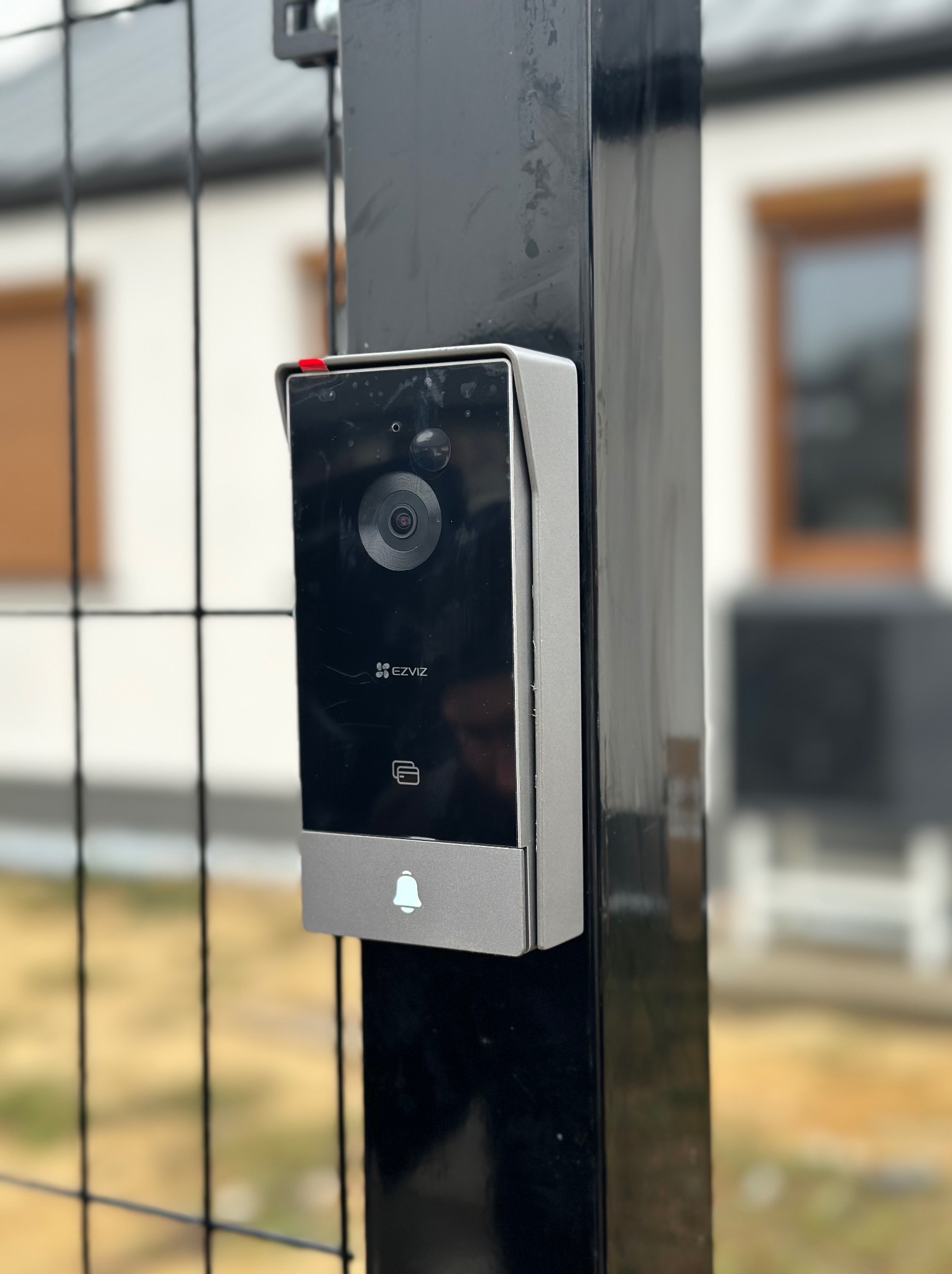 Nowoczesny wideodomofon marki EZVIZ zamontowany na czarnym słupku bramy, z widoczną kamerą, czytnikiem kart i przyciskiem dzwonka, na tle rozmytego budynku mieszkalnego i metalowej siatki...
