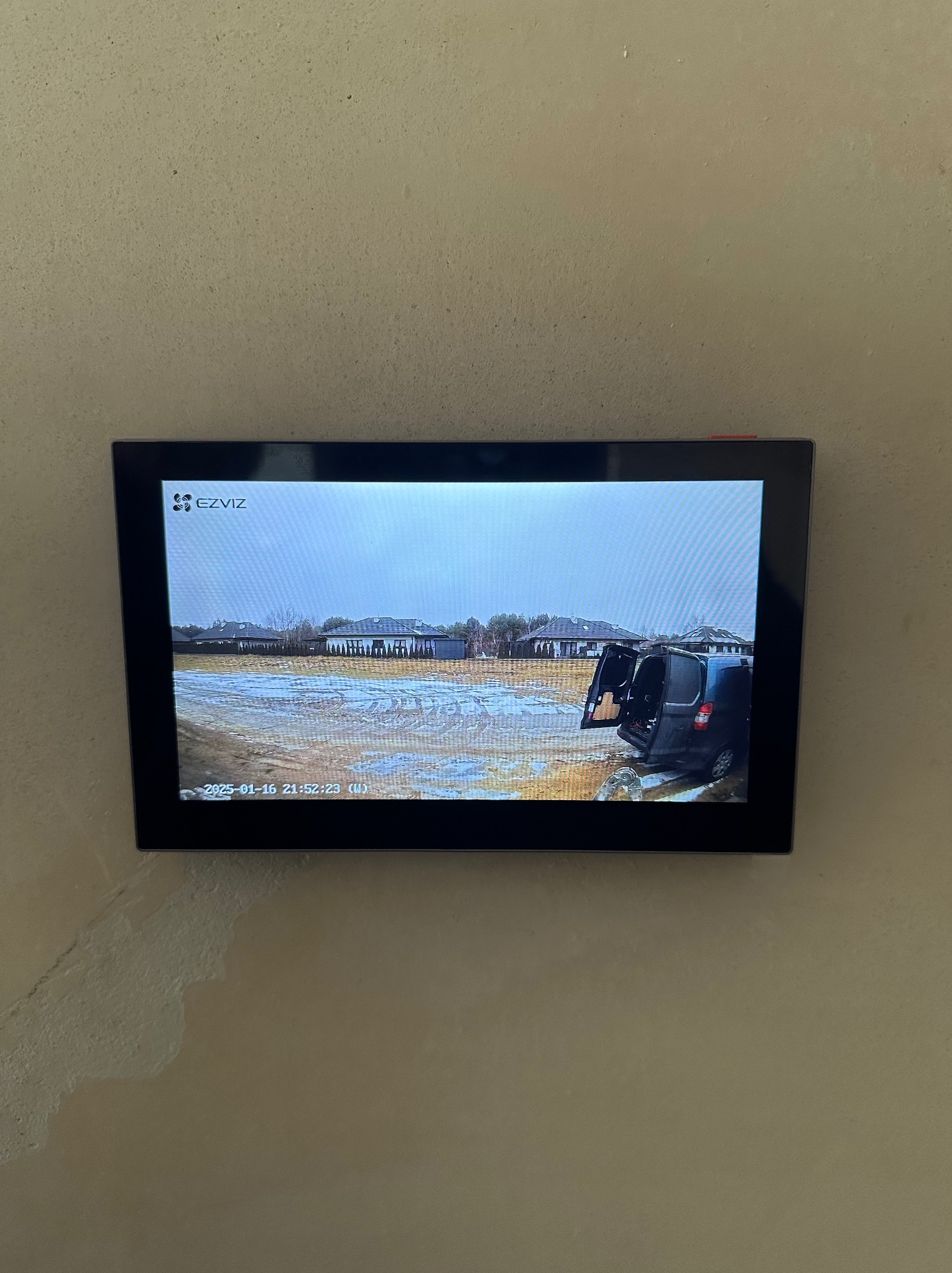 Ekran monitoringu na ścianie, prezentujący obraz z kamery: widoczny samochód dostawczy z otwartymi drzwiami w błotnistym terenie, domy w tle, data i godzina w lewym dolnym rogu.