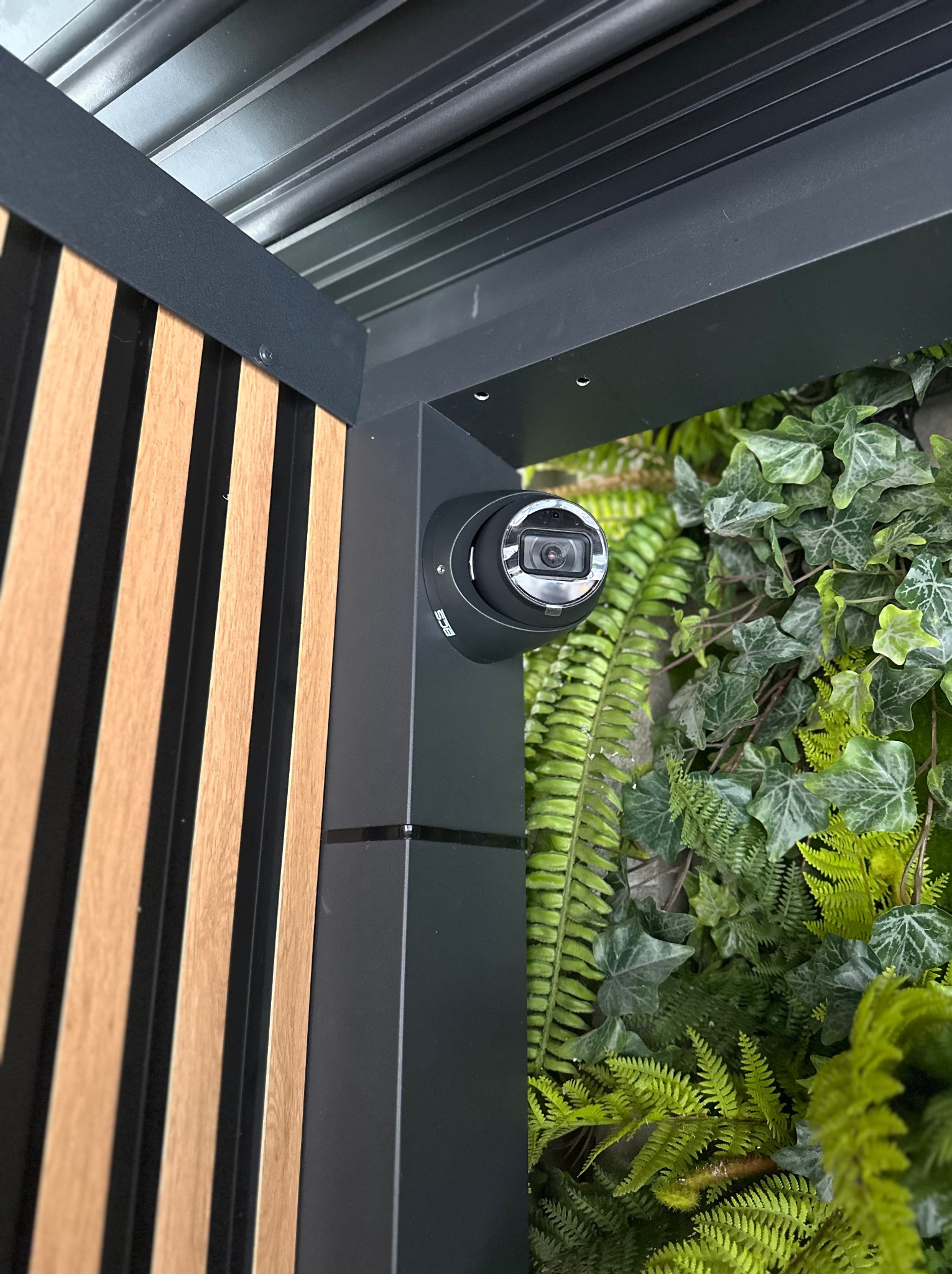 Kamera monitoringu zamontowana na czarnej kolumnie, obok drewnianych paneli i ściany z zielonych roślin.