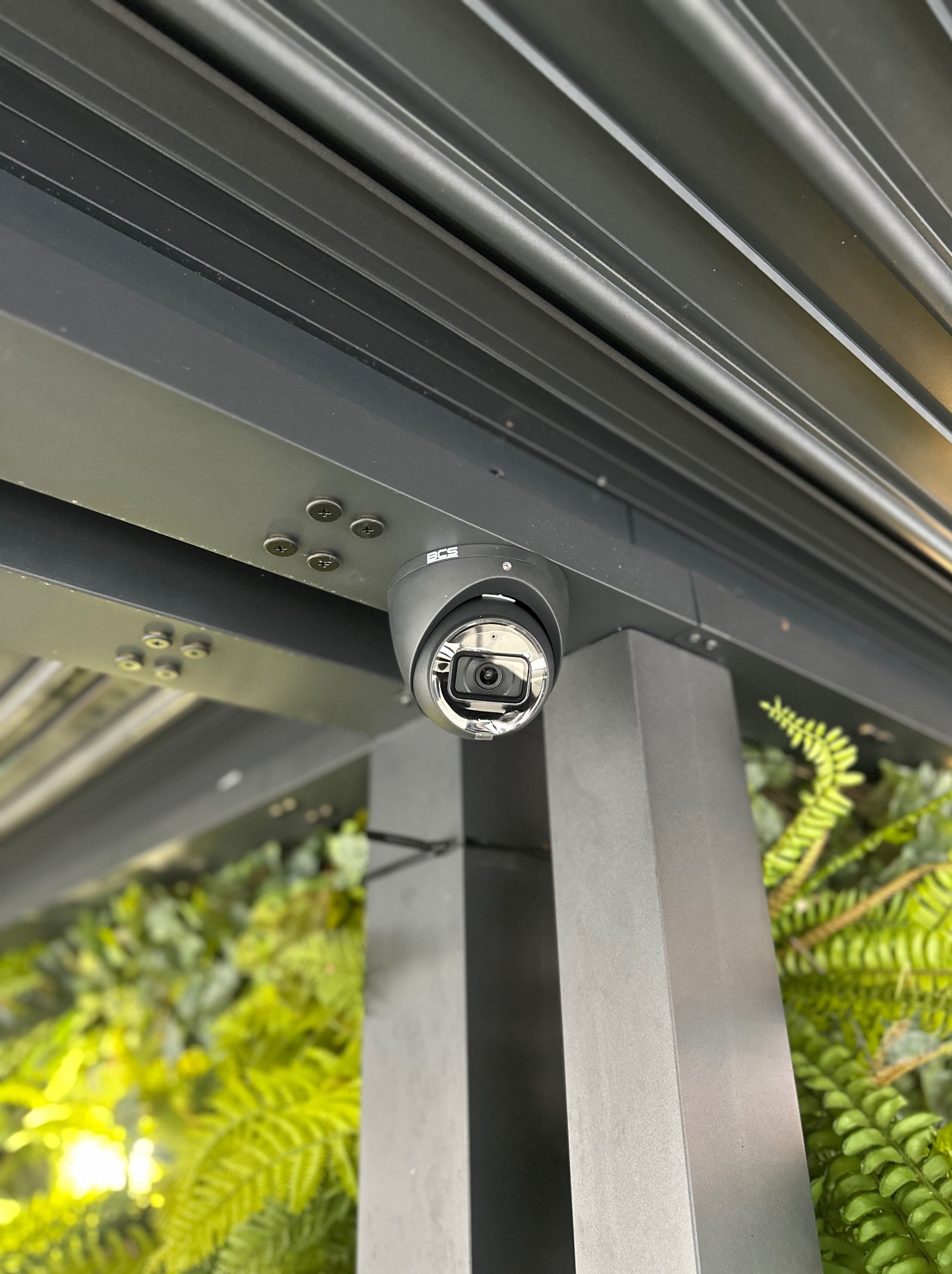 Kamera monitoringu kopułkowa, zamontowana pod nowoczesną, metalową konstrukcją z widocznymi śrubami, z zielonym tłem roślinnym.