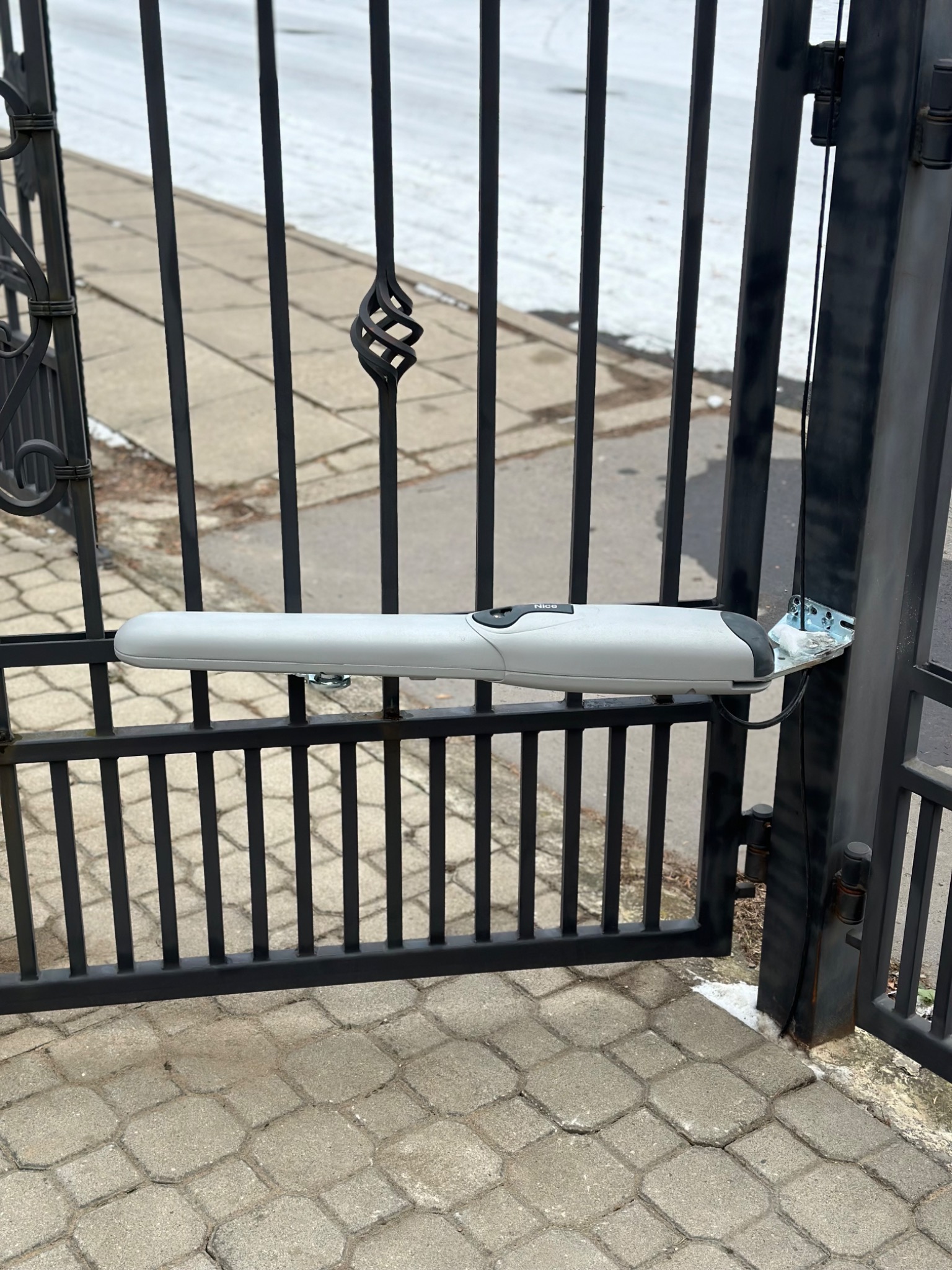 Zamontowany siłownik automatyki do bramy posesji w Olszewnicy Starej, widoczny na tle chodnika z kostki brukowej i resztek topniejącego śniegu.