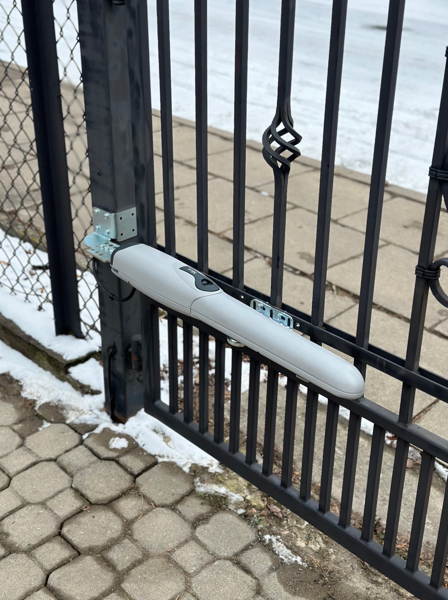 Szary siłownik bramy zamontowany na czarnej, metalowej bramie posesji. W tle widoczny chodnik z kostki i resztki śniegu.