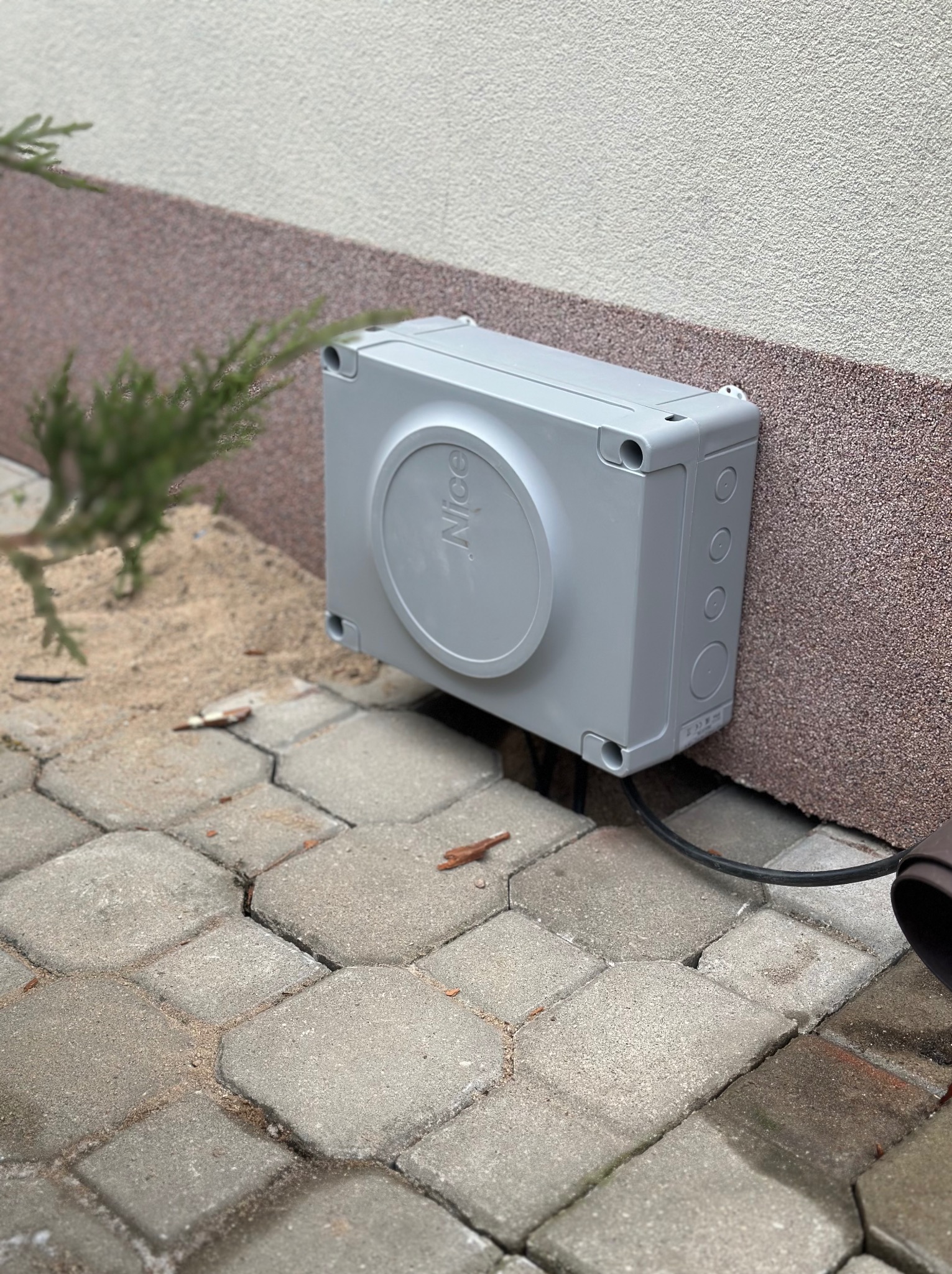 Szara skrzynka sterownicza z logo 'Nice' zamontowana na elewacji budynku, z widocznym kablem wychodzącym z dołu, obok kostki brukowej i małej rośliny.