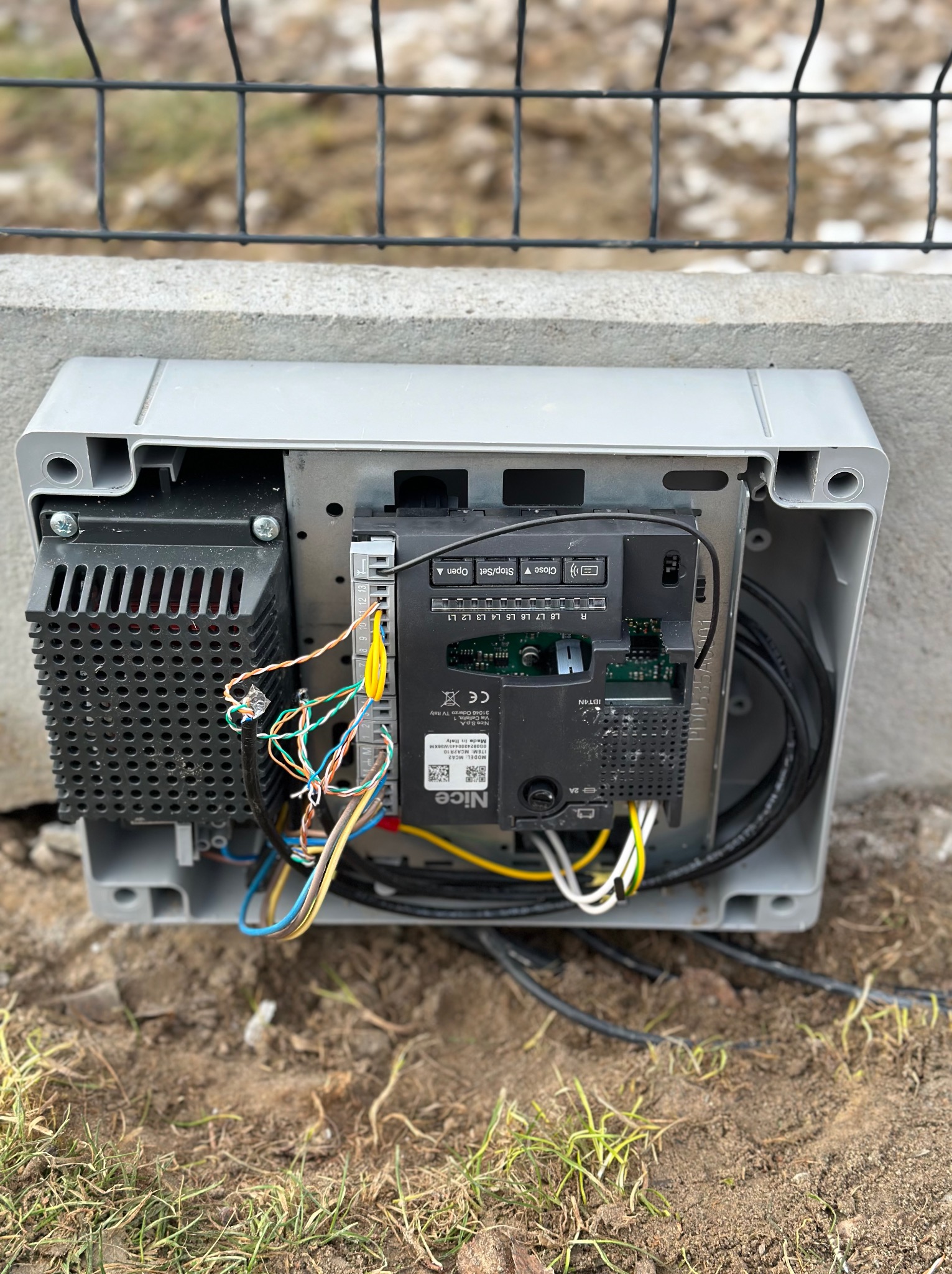 Otwarta obudowa sterownika bramy automatycznej Nice, widoczne okablowanie, moduł sterujący i radiator, zamontowana na betonowym fundamencie przy ogrodzeniu z siatki.
