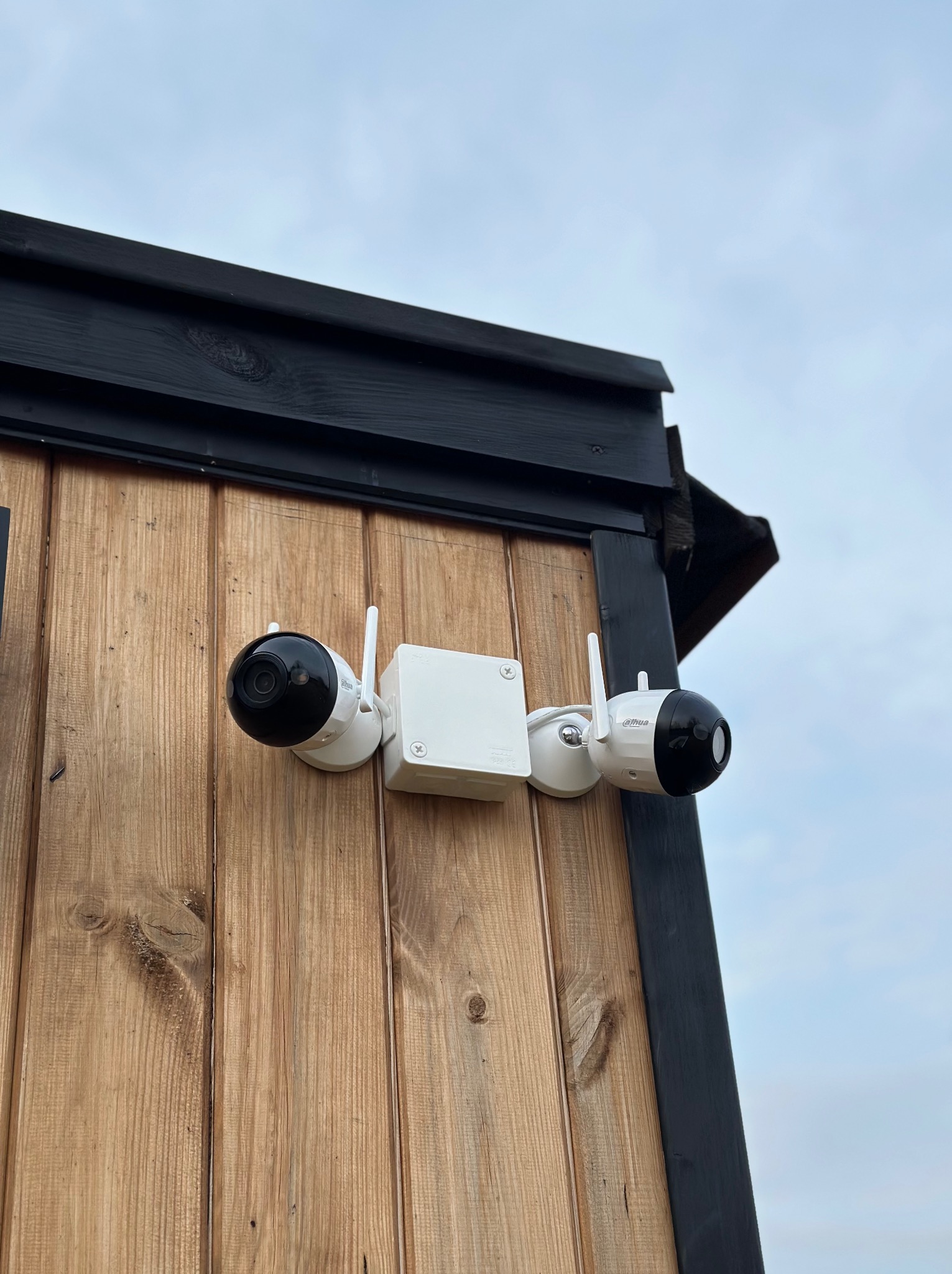 Dwie kamery monitoringu Dahua zamontowane na drewnianej ścianie budynku z czarnymi elementami wykończeniowymi, widok z dołu na tle pochmurnego nieba.