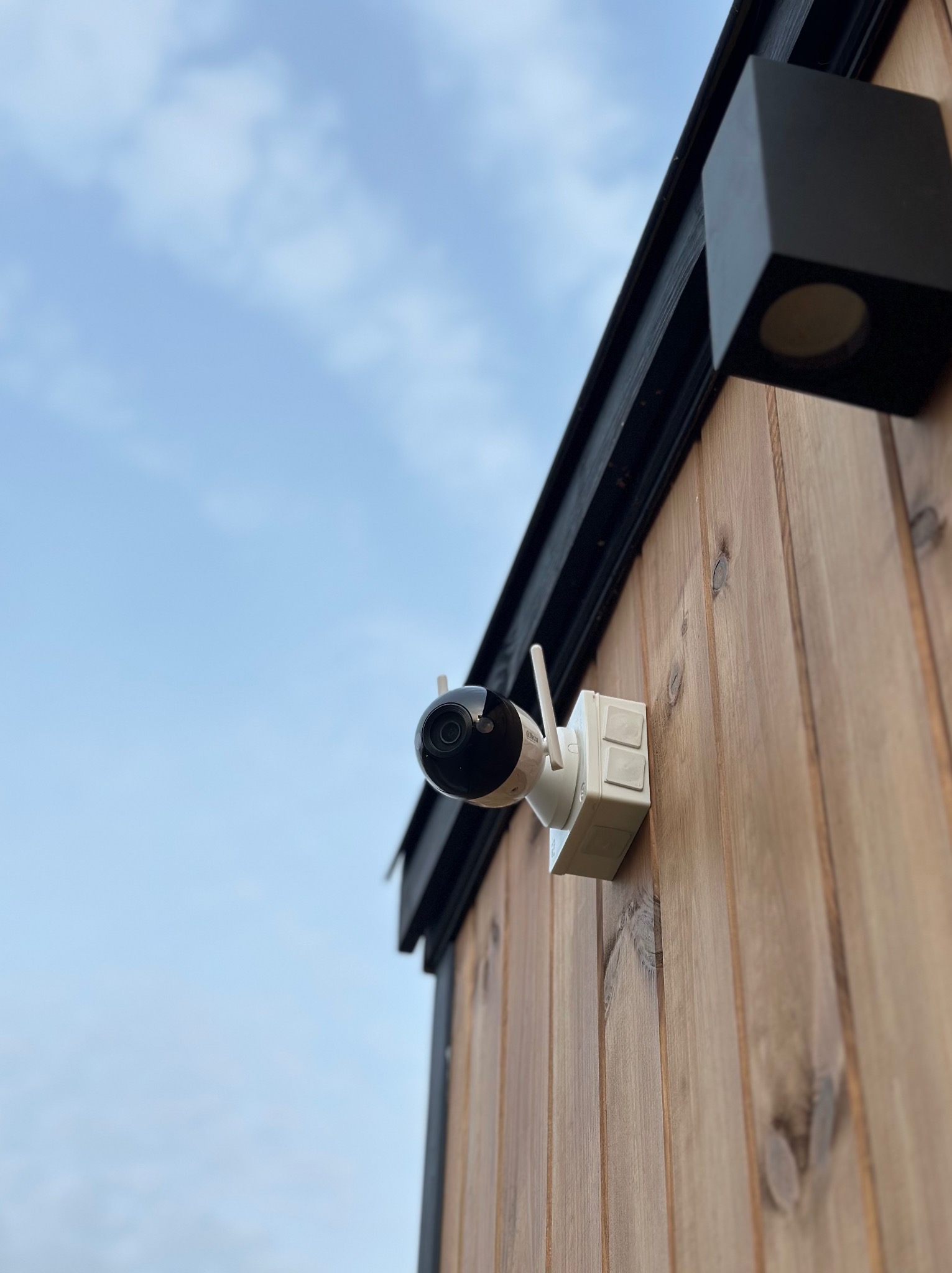Zewnętrzna kamera IP z antenami zamontowana na drewnianej elewacji budynku, obok czarnej lampy w kształcie sześcianu, na tle pochmurnego nieba.