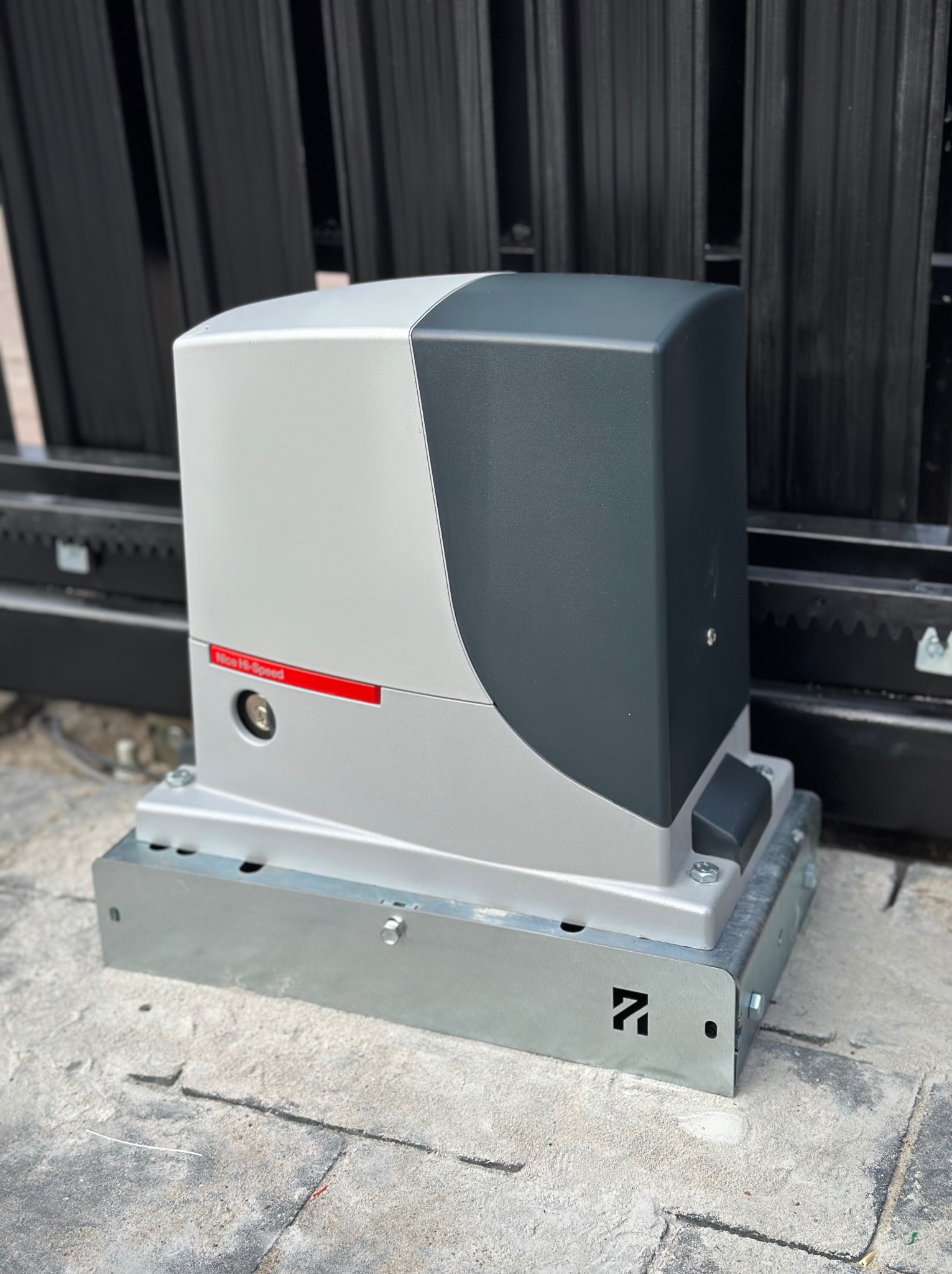 Zamknięty automat do bram przesuwnych Nice Hi-Speed w kolorze szaro-czarnym, zamontowany na metalowej podstawie, widoczny na tle czarnego ogrodzenia.