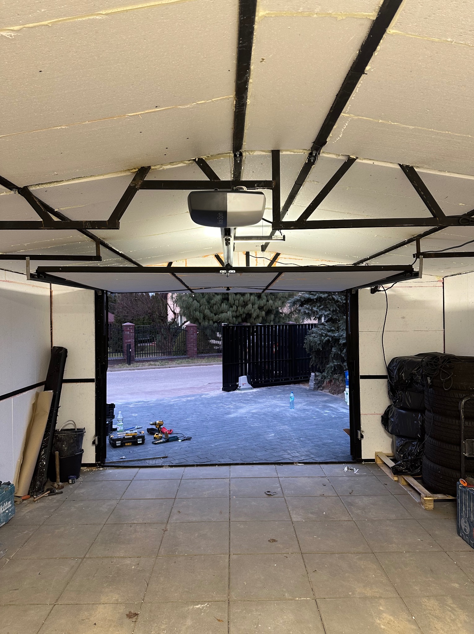 Wnętrze garażu z widocznym mechanizmem automatyki bramy segmentowej podczas montażu, narzędzia rozłożone na zewnątrz, podłoga wyłożona płytkami.