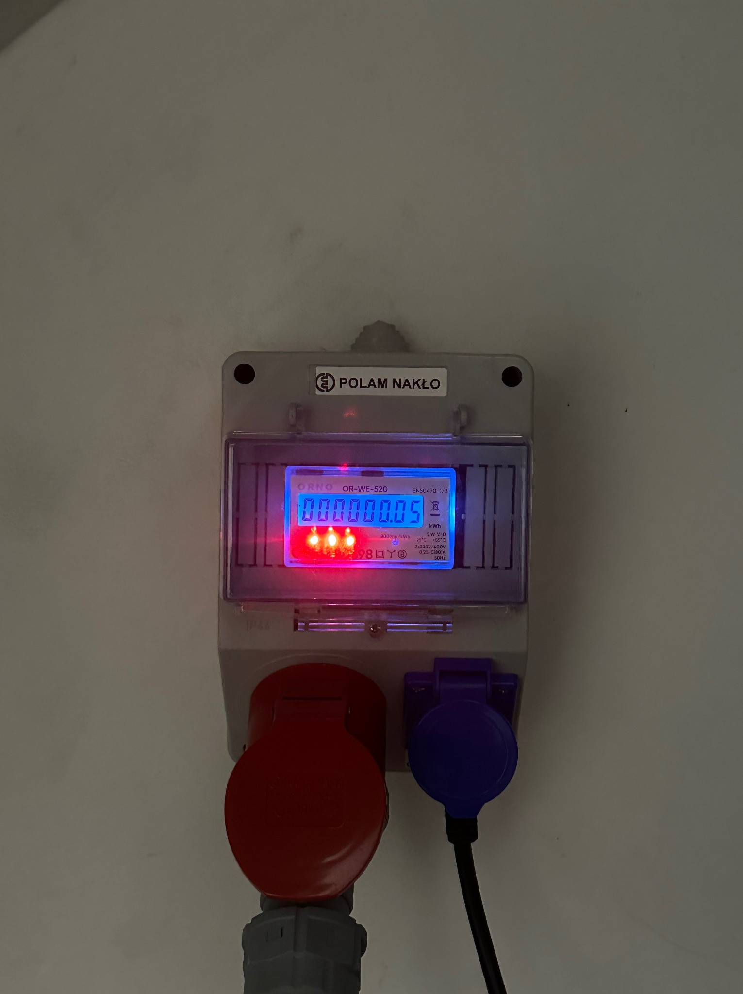 Szary licznik energii elektrycznej ORNO OR-WE-520 z czerwonym i niebieskim gniazdem na białej ścianie. Wyświetlacz pokazuje zużycie energii.