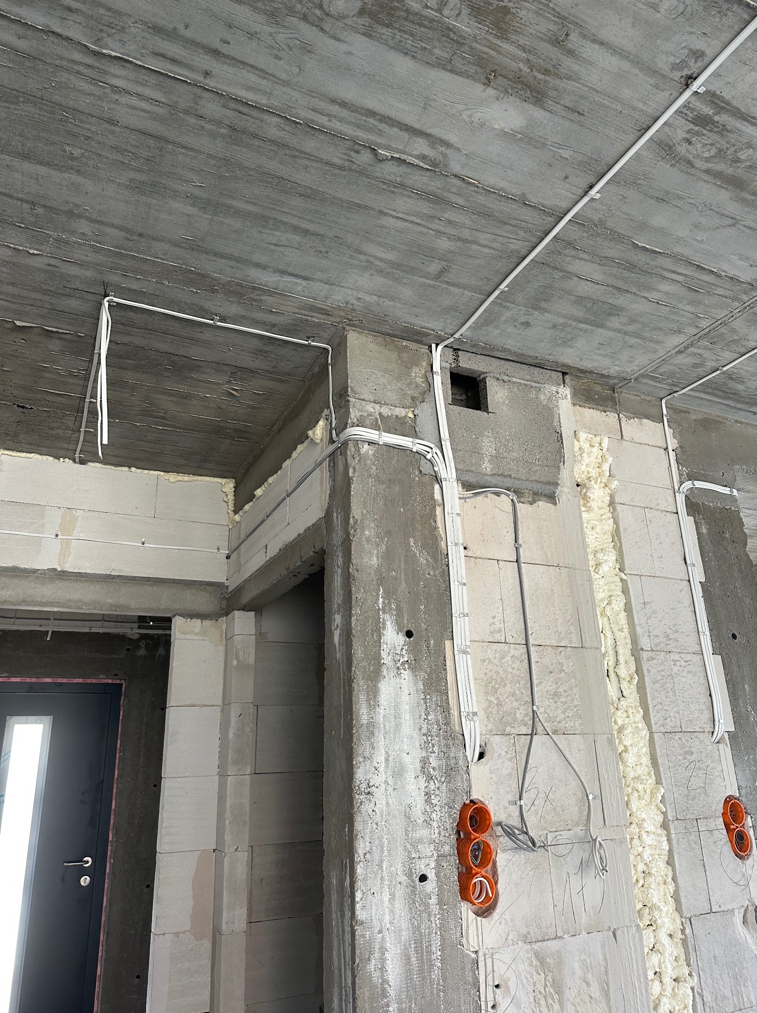 Surowy stan: instalacja elektryczna w budynku z betonu komórkowego, widoczne kable w peszlach, puszki elektryczne i pianka montażowa. Szare ściany i sufit.