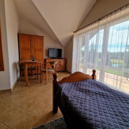 FACELIA SPÓŁKA Z OGRANICZONĄ ODPOWIEDZIALNOŚCIĄ - Pokój hotelowy na poddaszu z drewnianym łóżkiem przykrytym granatową narzutą, widokiem na jezioro przez duże okno z białymi firankami, drewnianymi meblami i lustrem.