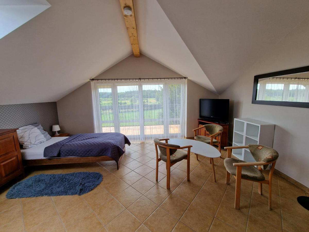 Pokój hotelowy na poddaszu z łóżkiem, stolikiem kawowym i widokiem na zielone pola przez okno balkonowe z firankami.