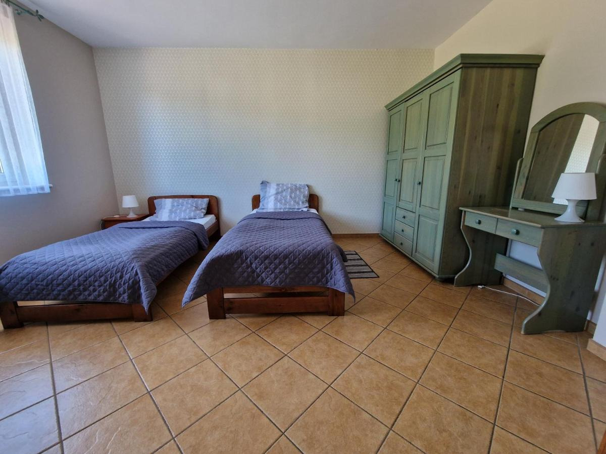 Pokój dwuosobowy z dwoma łóżkami pojedynczymi z drewnianymi ramami i szarymi narzutami, zielona szafa i toaletka z lustrem w tle, brązowe płytki na podłodze.