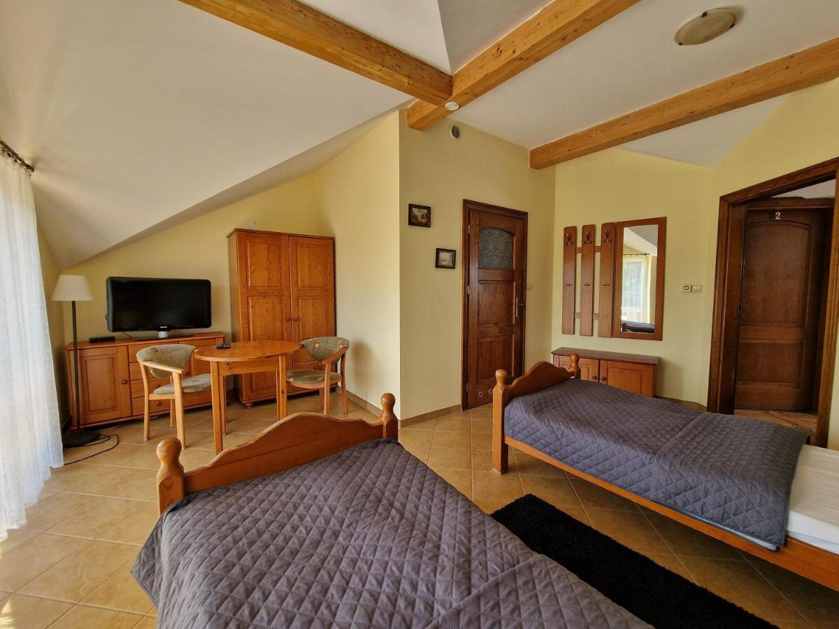 Pokój dwuosobowy z drewnianymi łóżkami, stolikiem i telewizorem, utrzymany w ciepłej, żółtej kolorystyce z drewnianymi elementami wykończeniowymi.