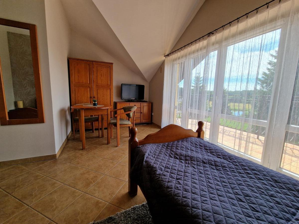 Pokój hotelowy na poddaszu z drewnianym łóżkiem przykrytym granatową narzutą, widokiem na jezioro przez duże okno z białymi firankami, drewnianymi meblami i lustrem.