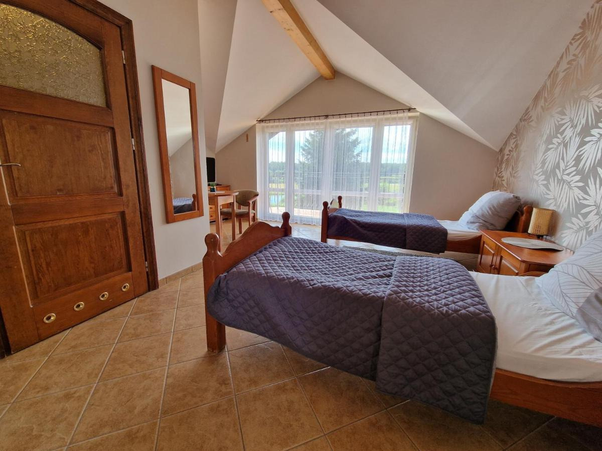 Pokój dwuosobowy na poddaszu z dwoma pojedynczymi łóżkami z drewnianymi ramami i szarymi pikowanymi narzutami, widok na zewnątrz przez okno z firankami, wzorzysta tapeta na ścianie.