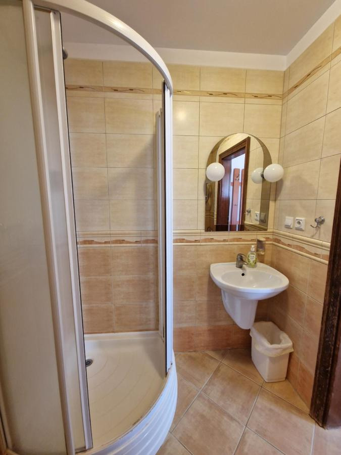 Łazienka z kabiną prysznicową, umywalką i lustrem w stylu retro, wykończona beżowymi płytkami ceramicznymi z dekoracyjnym pasem. Widoczny kosz na śmieci i butelka mydła.