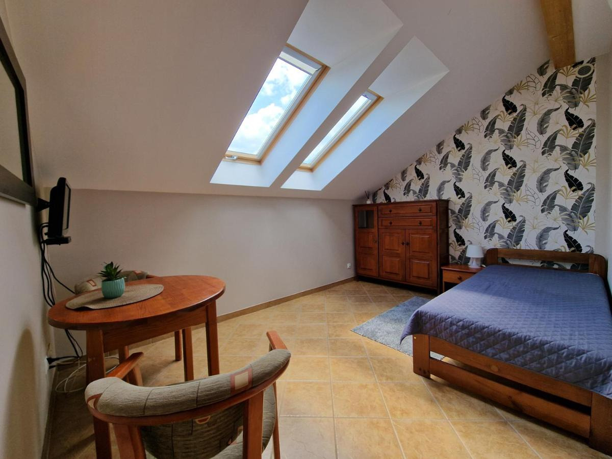 Pokój na poddaszu z drewnianym łóżkiem przy ścianie w liście, okrągłym stołem z rośliną w doniczce i trzema oknami dachowymi w skosie.