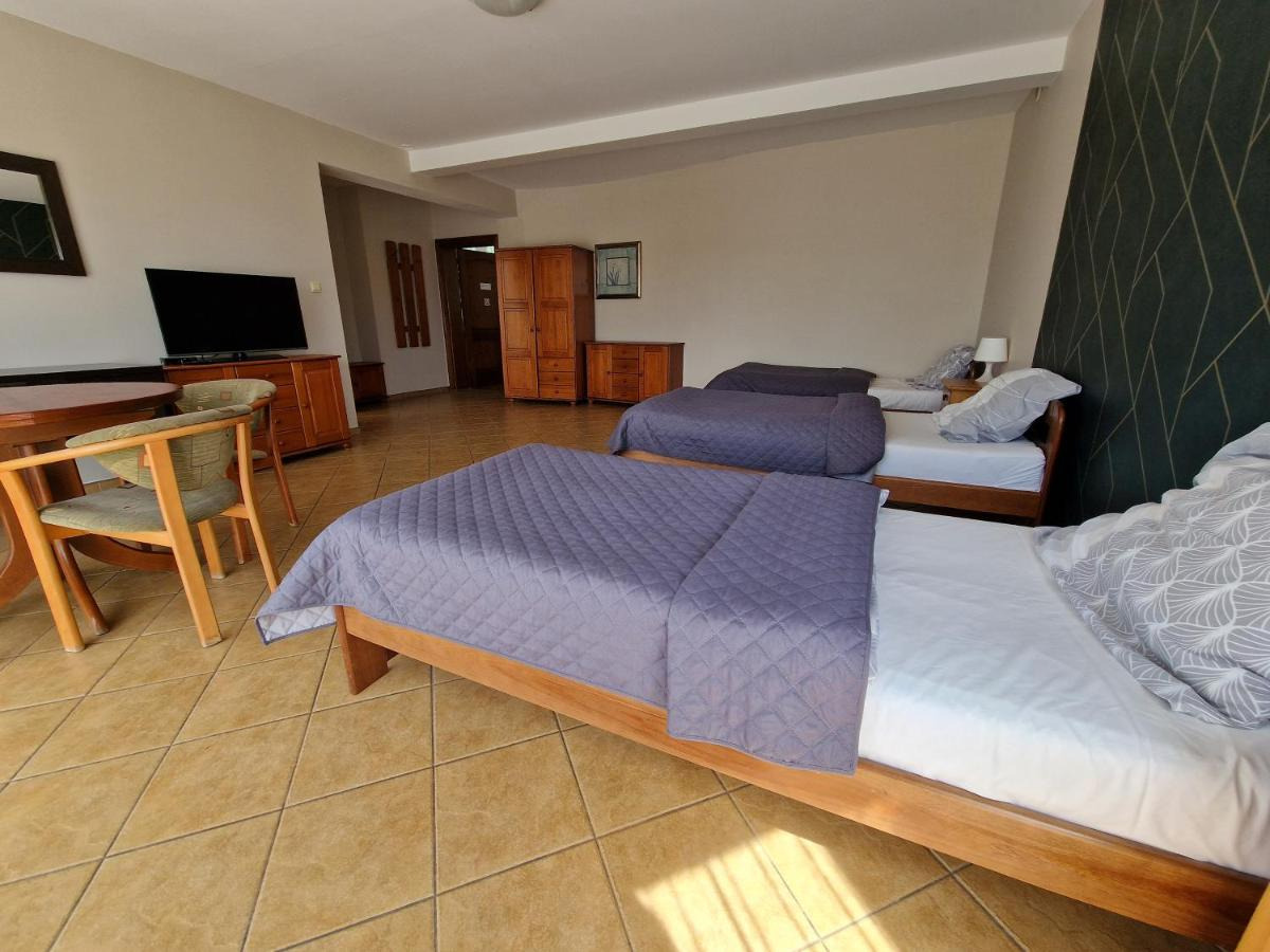 Pokój hotelowy z trzema pojedynczymi łóżkami z drewnianymi ramami i fioletowymi narzutami, drewniane meble, podłoga z płytek, ciemna ściana z geometrycznym wzorem.