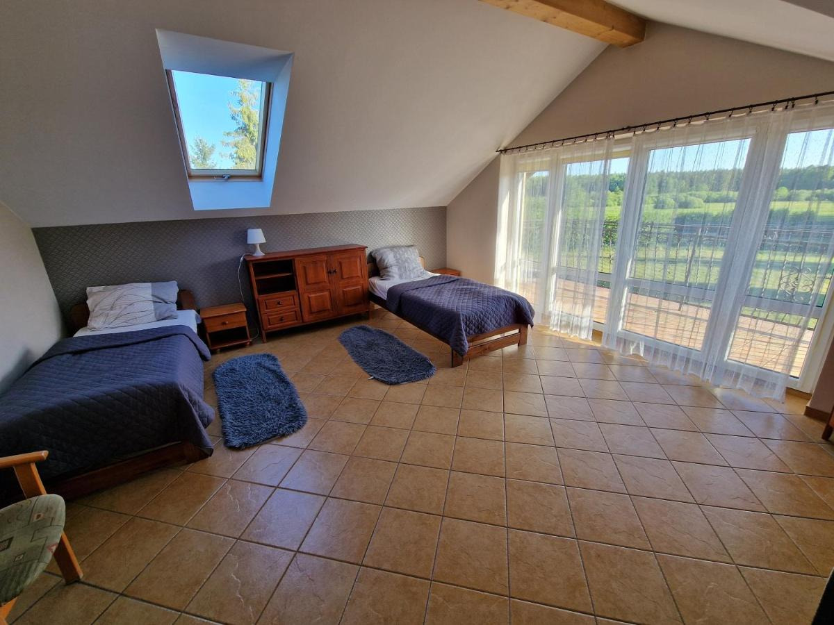 Pokój na poddaszu z dwoma pojedynczymi łóżkami, drewnianą komodą, oknem dachowym i dużymi oknami balkonowymi z widokiem na zieleń.