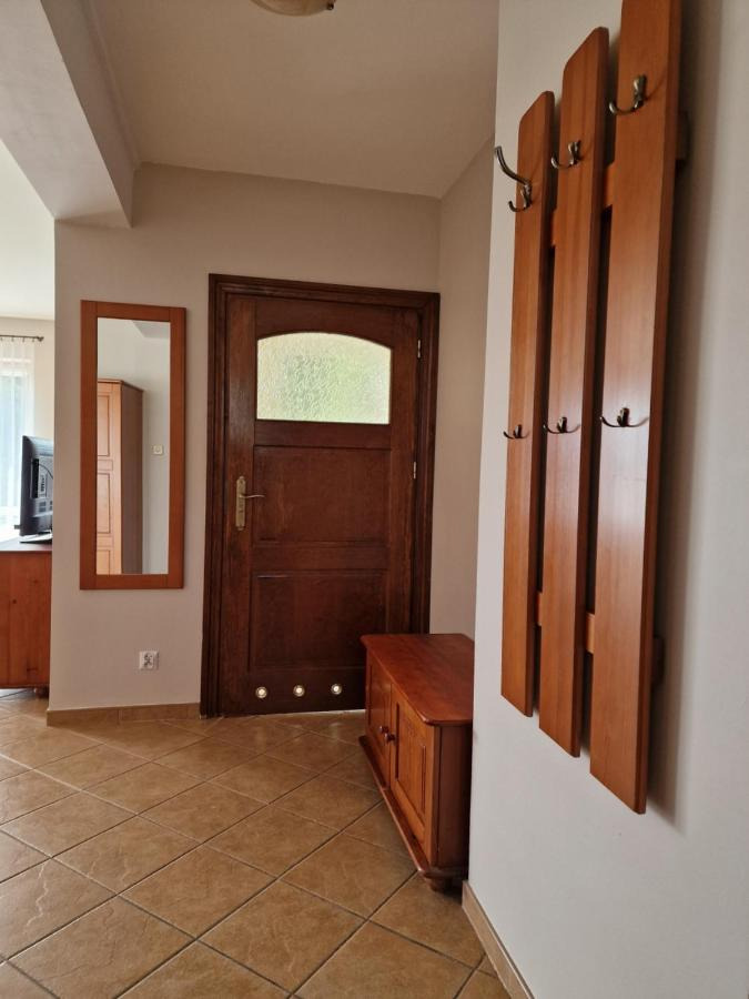 Drewniany przedpokój z ciemnymi płytkami podłogowymi, lustrem w ramie, wieszakiem z haczykami i niską komodą przy drzwiach.