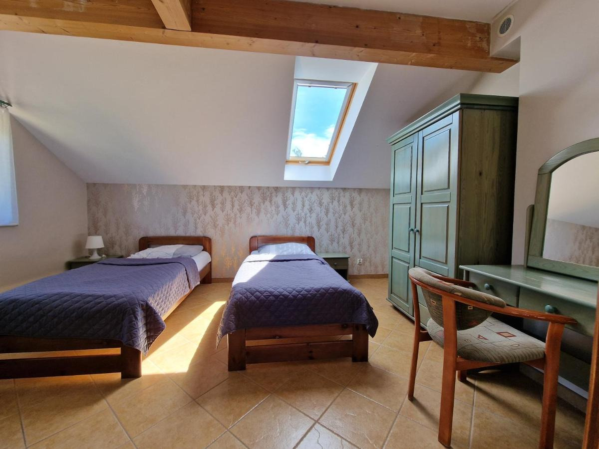 Pokój na poddaszu z dwoma łóżkami pojedynczymi z drewnianymi ramami, niebieskimi narzutami, oknem dachowym i zieloną szafą oraz toaletką, oświetlony naturalnym światłem.