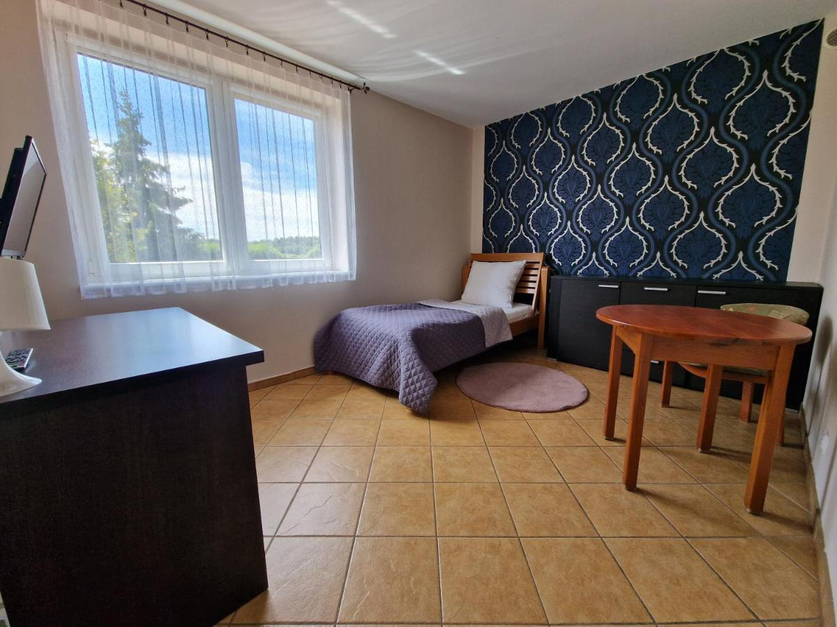 Pokój z pojedynczym łóżkiem przy oknie z widokiem na zieleń, wzorzysta tapeta na jednej ścianie, drewniany stół i krzesła, brązowe płytki podłogowe.