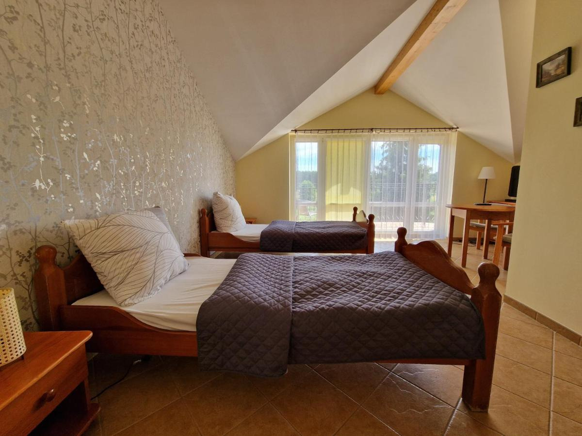 Pokój w hotelu SPA na poddaszu z dwoma pojedynczymi łóżkami z drewnianymi ramami i szarymi narzutami, okno z widokiem na zieleń, drewniany stół z lampką.