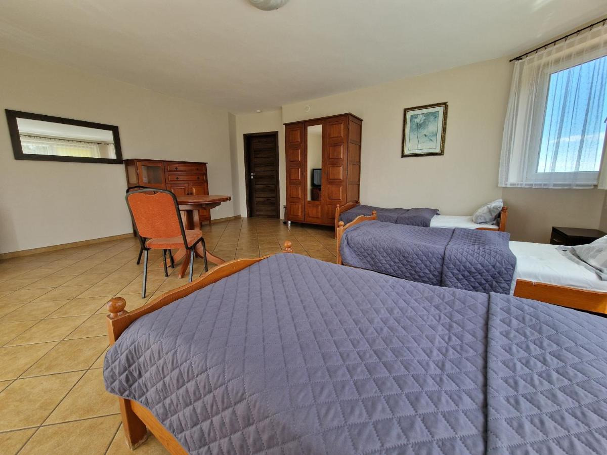 Pokój z trzema łóżkami pojedynczymi z szarymi narzutami w kratę, drewnianą szafą z lustrem, komodą i stołem z krzesłem, jasne ściany, okno z białymi firankami.