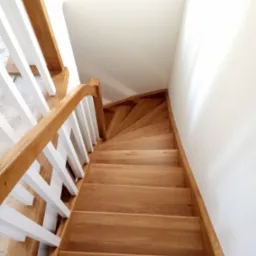 Kątowe ujęcie drewnianych schodów z białymi balustradami, prowadzących w górę w jasnym wnętrzu.