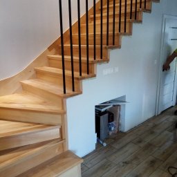 Zakład Stolarski Paweł Talaśka - Jasne, drewniane schody z czarnymi, pionowymi poręczami, częściowo zamontowane, z widocznym schowkiem pod spodem i fragmentem osoby po prawej stronie.