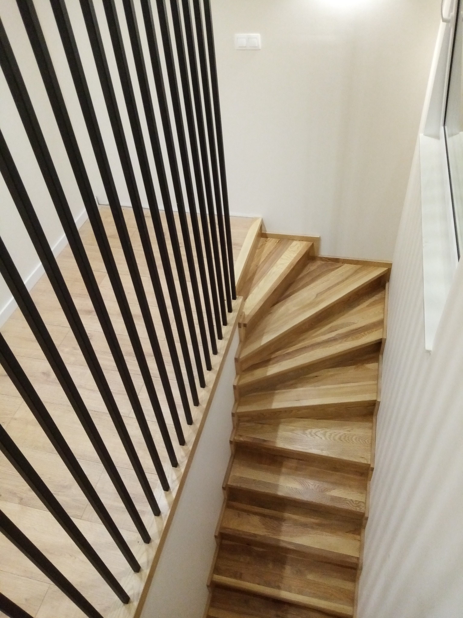 Drewniane schody spiralne z jasnego drewna z czarną, metalową balustradą pionową, widok z góry, oświetlenie naturalne.