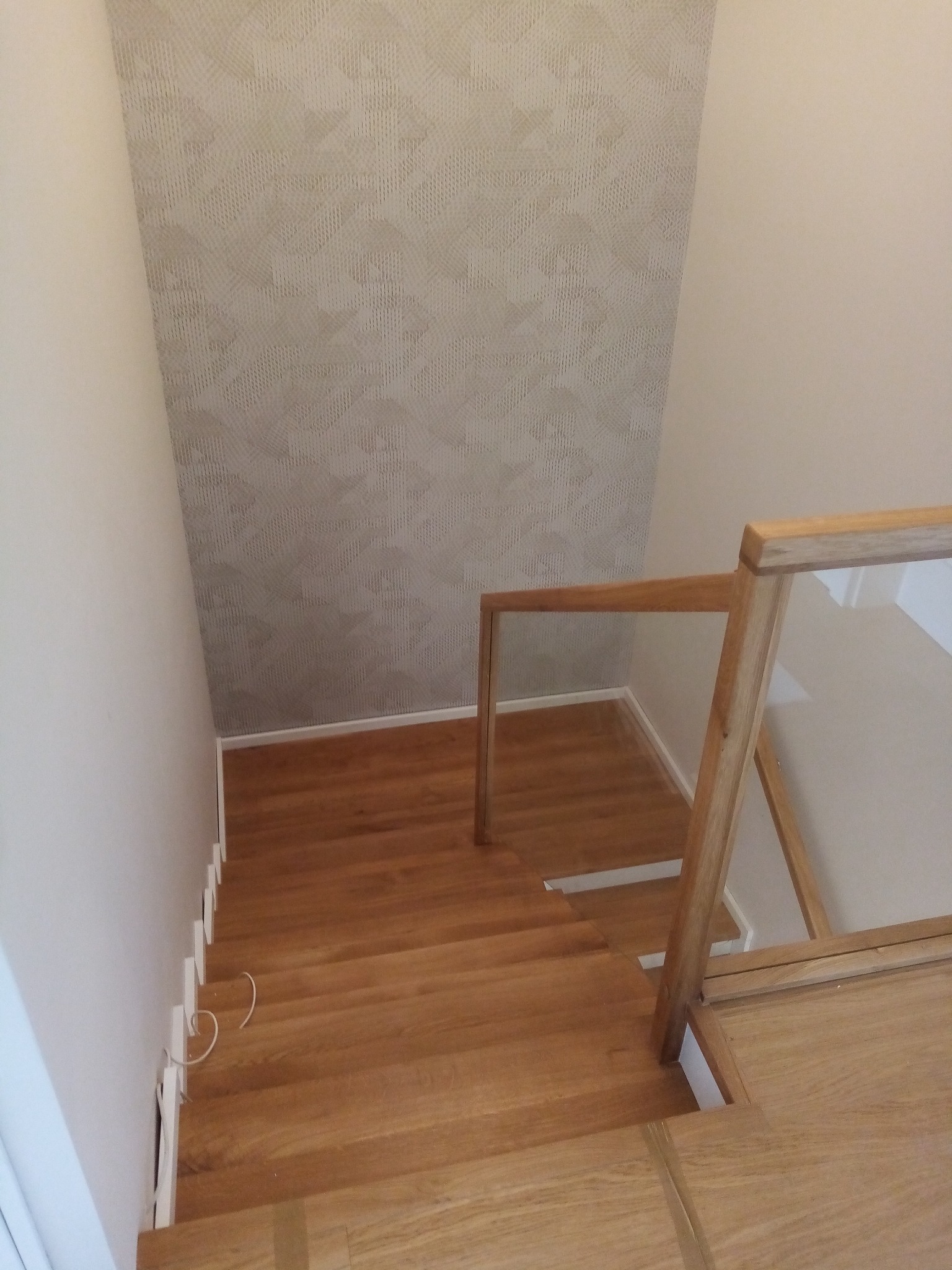 Widok z góry na jasne, drewniane schody w Śliwicach z nowoczesną balustradą ze szkła i drewna; ściana pokryta tapetą w geometryczny wzór.