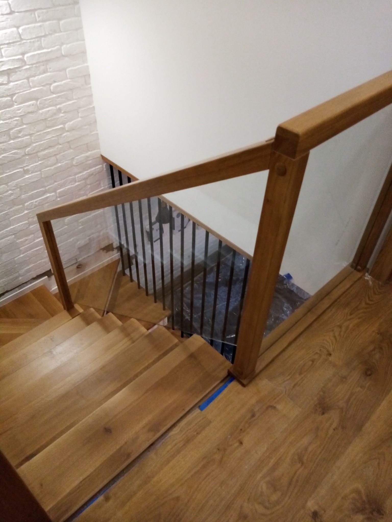 Klatka schodowa z drewnianymi stopniami i szklaną balustradą z drewnianą ramą, widok z góry, ściana z białej cegły w tle.